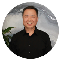 A man with short black hair is smiling, wearing a black shirt, and standing next to a plant. The image is circular.