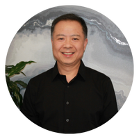 A man with short black hair is smiling, wearing a black shirt, and standing next to a plant. The image is circular.