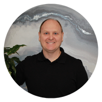 A man is smiling, wearing a black shirt, and standing next to a plant with a soft background. The image is circular.
