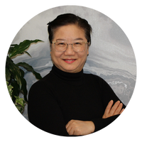 A woman with short hair is smiling and standing with her arms crossed, wearing a black turtleneck shirt. The image is circular.