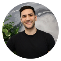A man with short dark hair is smiling, wearing a black shirt, and standing next to a plant. The image is circular.