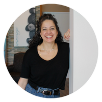 A woman with curly hair is smiling and leaning against a doorframe, wearing a black shirt and jeans. The image is circular.