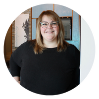 A woman with glasses and blonde hair is smiling, wearing a black shirt, and standing in front of a wall with artwork. The image is circular.