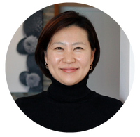 A woman with short dark hair is smiling, wearing a black turtleneck shirt. The image is circular.