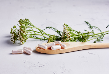 A wooden spoon with several capsules resting on it, alongside fresh green herbs and flowers. The background is light and neutral.