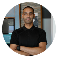 A man with short hair is standing with his arms crossed, wearing a black shirt. The image is circular.