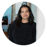 A woman with long dark hair is standing, wearing a black sweater. The image is circular.