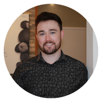 A man with a beard is smiling, wearing a patterned black shirt. The image is circular.