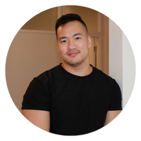 A man with short hair is smiling, wearing a black shirt, and standing next to a doorframe. The image is circular.
