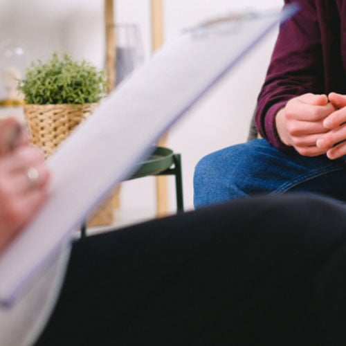 A close-up of two people in a therapy or counseling session. One person is holding a clipboard while the other is seated, with their hands visible. The background features a plant and a cozy, calming environment.